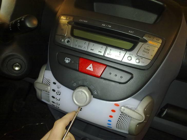 Sådan afmonteres radio i Aygo - Løft forsigtigt knappen af med en skruetrækker. Brug gerne en plastik spartel eller ligende hvis du har, for at være sikker på at undgå mærker. billede 1