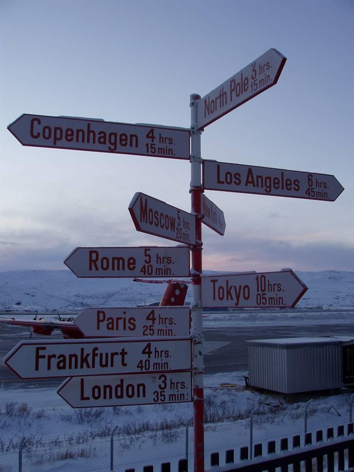 Greenland - Grønland - Kalaallit Nunaat (1) - Tilbage i Kangerlussuaq´s lufthavn. Udenfor står et skilt, som viser hvor mange timer det ville tage at flyve diverse steder hen, samt i hvilken retning de ligger, hvis der altså ikke kun fløj fly derfra til Danmark. Man kan lige ane New York (city) i baggrunden, som det kun ville tage ca. 4 timer og 0 minutter at flyve til. billede 15